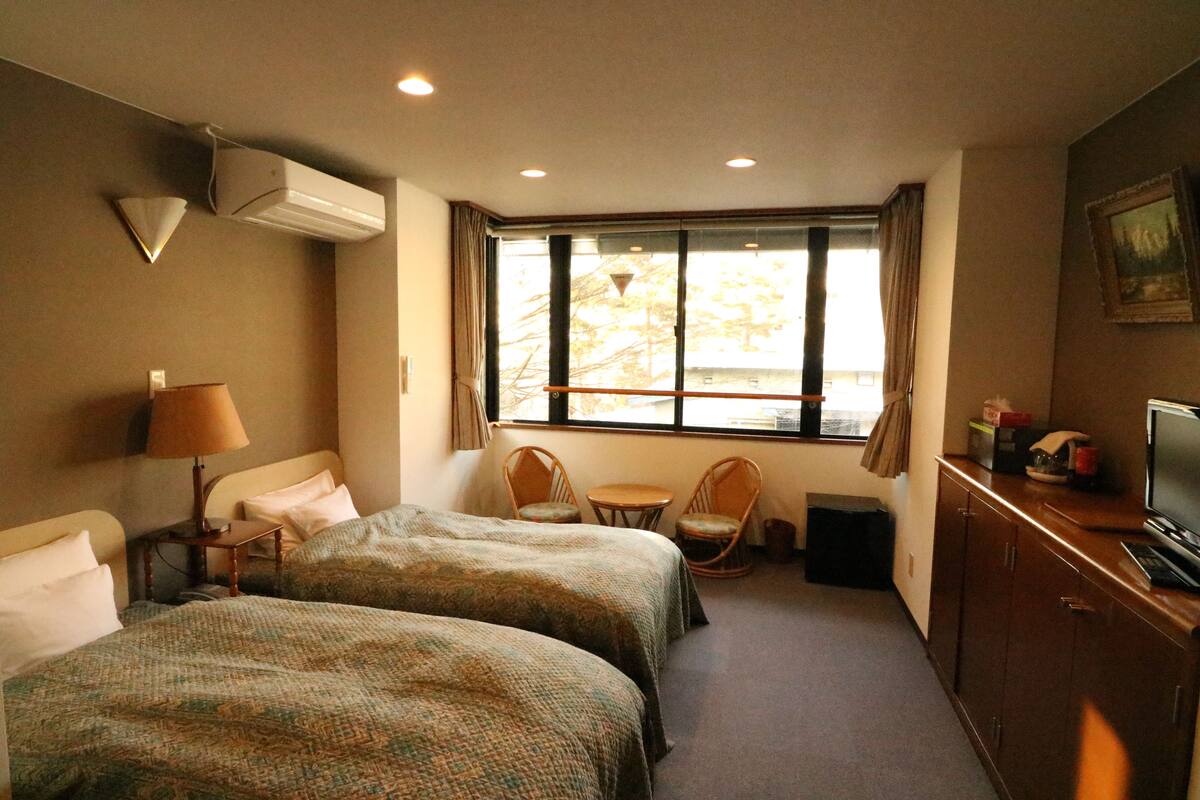 A hotel room with two beds, a small table, two chairs, a TV, and a window with a view of trees.