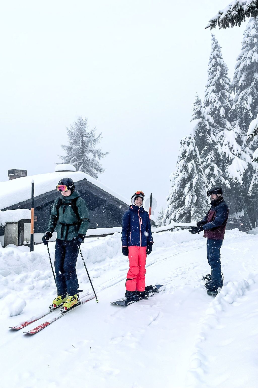 Trois personnes font du ski dans un paysage enneigé, avec des arbres et un bâtiment en arrière-plan.