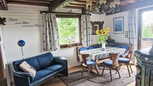Ein gemütliches Zimmer mit einem blauen Sofa, einem Holzesstisch und Stühlen. Eine Chandelüster, ein Fenster mit Vorhängen und ein Gemälde schmücken die Wand.