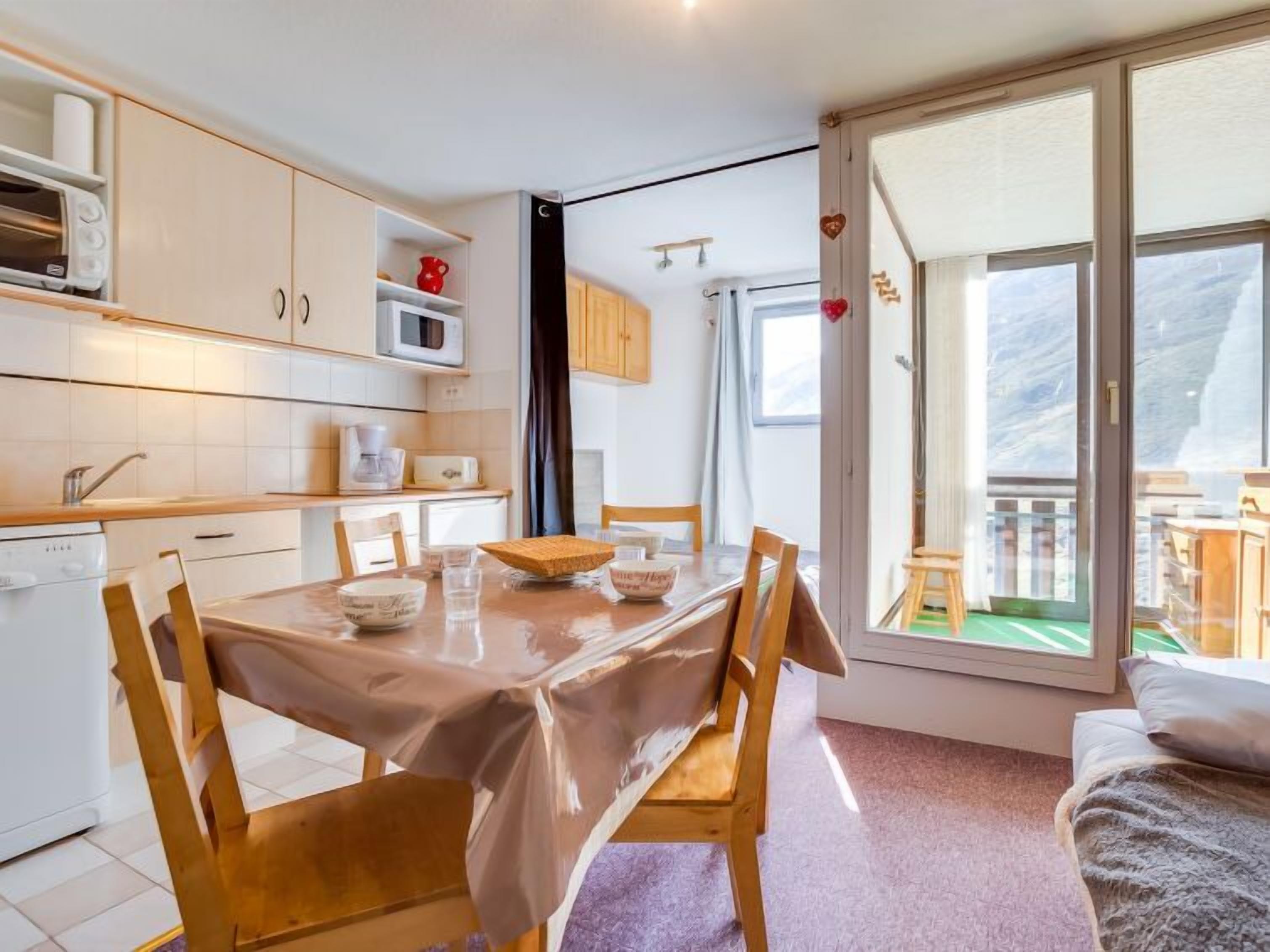Cocina compacta con muebles blancos, microondas y cafetera. Hay una mesa de comedor con sillas, un taburete pequeño y una cama con manta.