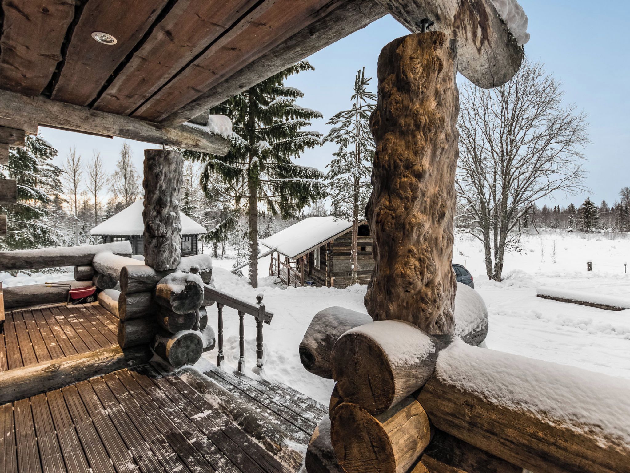 A wooden cabin with a snow-covered deck, logs stacked outside, and a clear view of the surrounding snowy landscape.