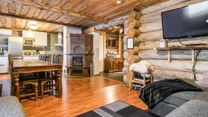 Ein gemütliches Blockhaus-Interieur mit einem Holztisch, Kamin, Fernseher an einer Baumstammmauer und einem durch eine offene Tür erreichbaren Küchenbereich.