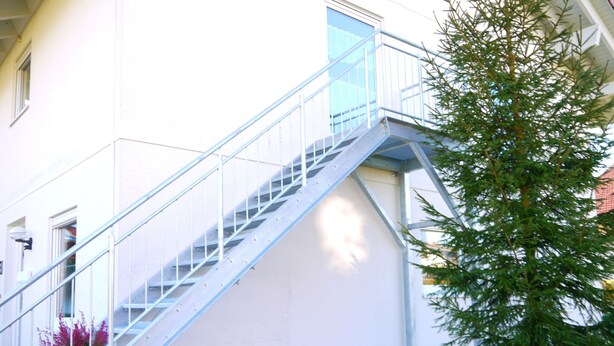A building with a metal staircase leading to a balcony, surrounded by a gravel area and a small tree.
