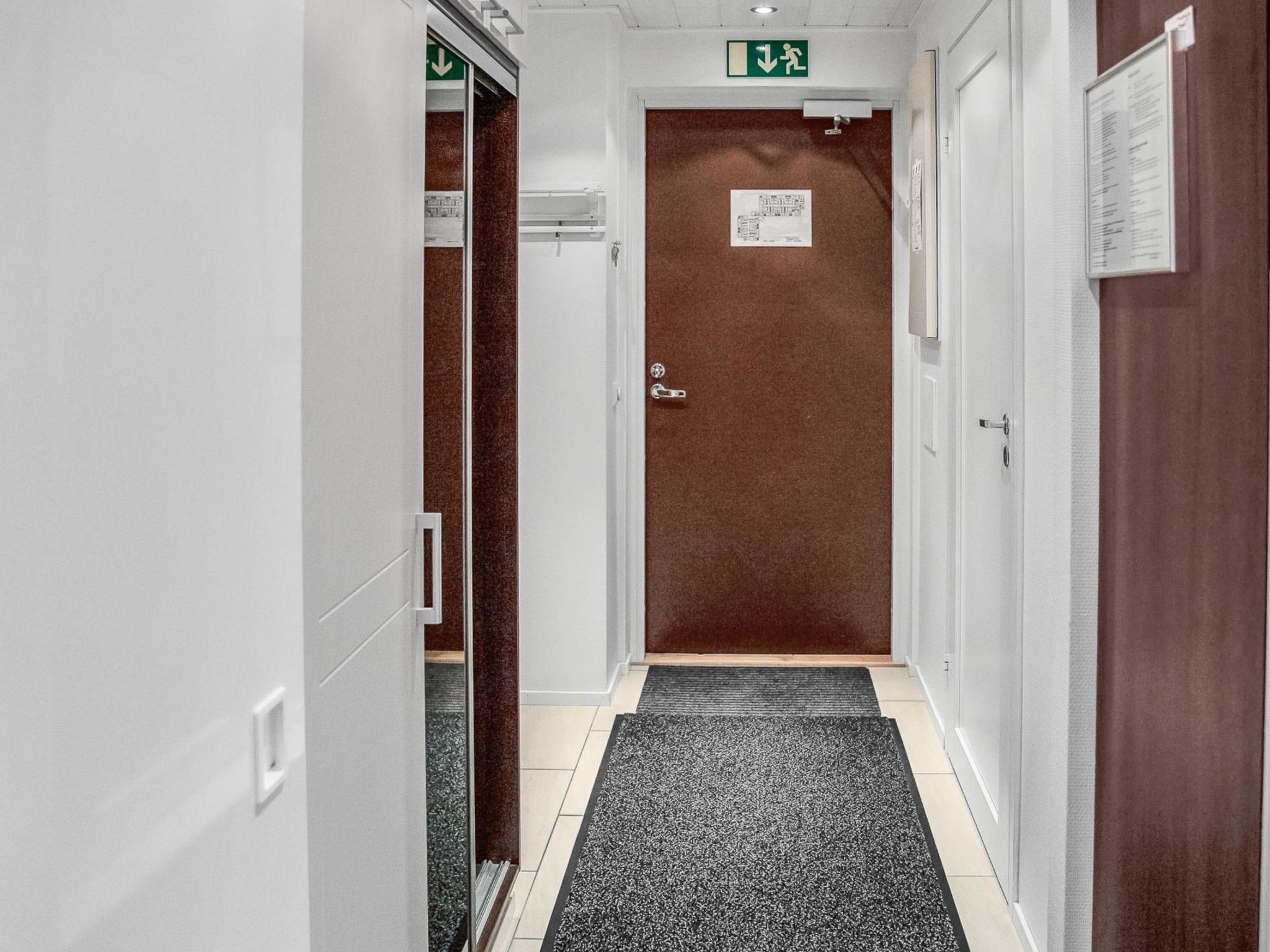 Un pasillo con una puerta marrón, un letrero de salida verde y una alfombra negra y gris.