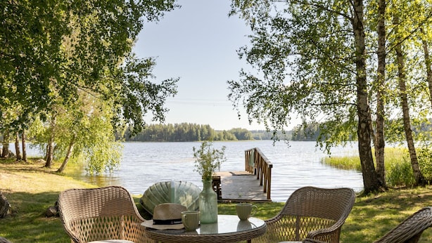 Ein Sitzbereich im Freien mit Korbstühlen und einem Tisch, mit Blick auf einen See mit einem Steg.