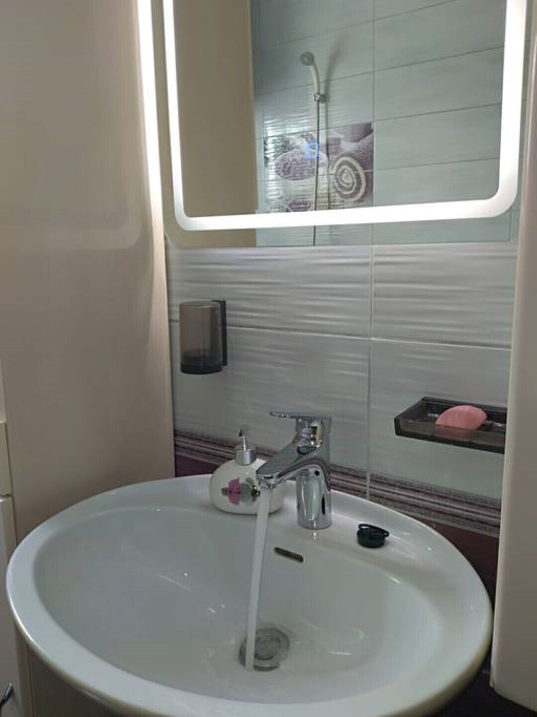 Ein Badezimmer mit einem weißen Waschbecken, einem Spiegel darüber und einer in der Spiegelung sichtbaren Dusche.