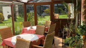 A covered patio area with wooden tables and chairs, large windows, and potted plants.