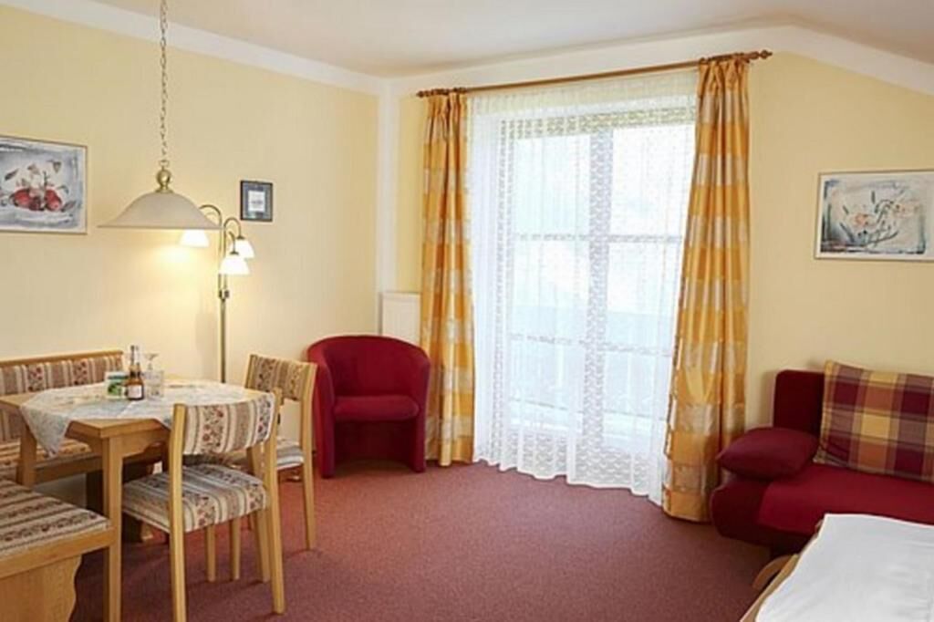 A room with a dining table, chairs, a red armchair, a sofa, and a window with curtains.