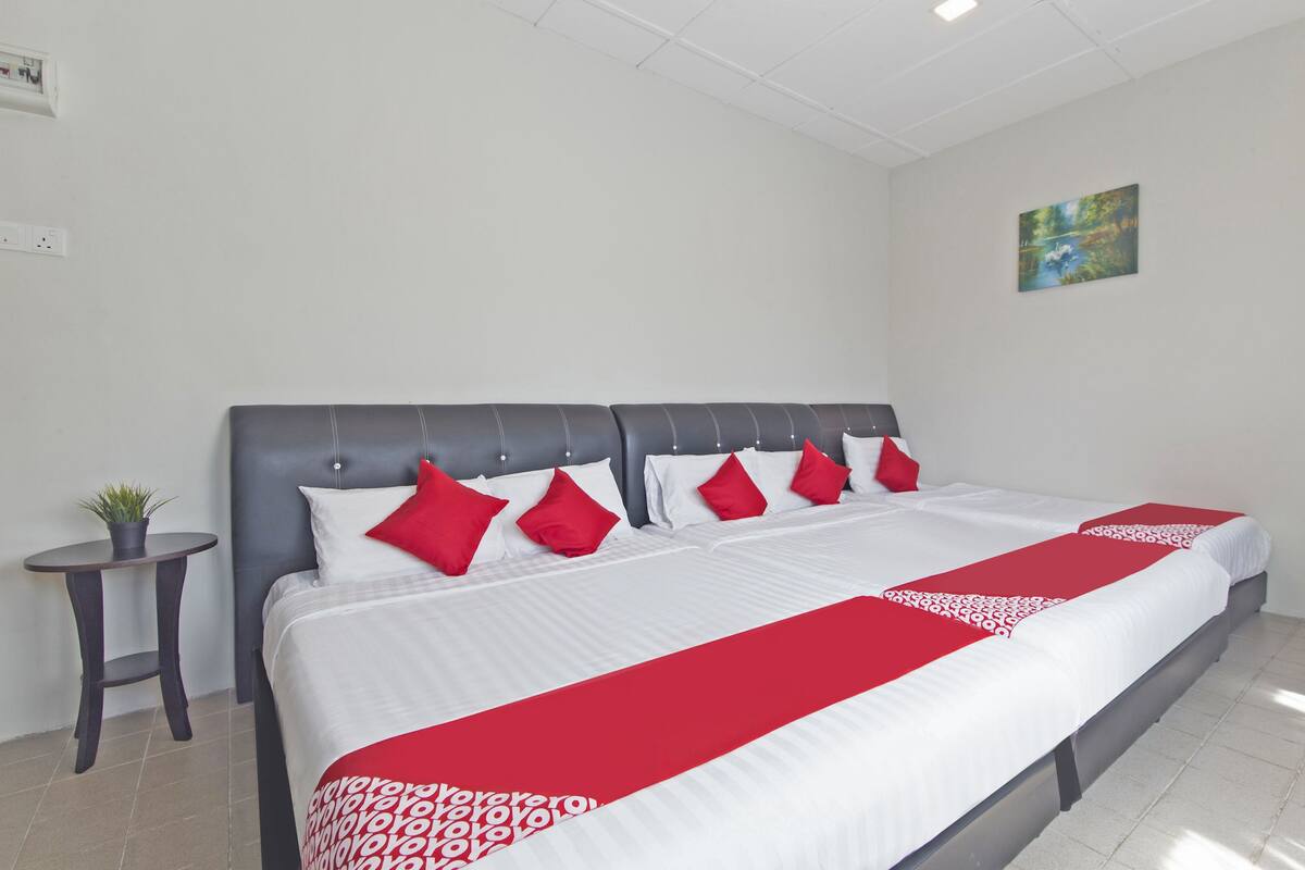 Una habitación de hotel con una cama grande, ropa de cama blanca, detalles en rojo, una mesita de noche con una planta y un cuadro en la pared.