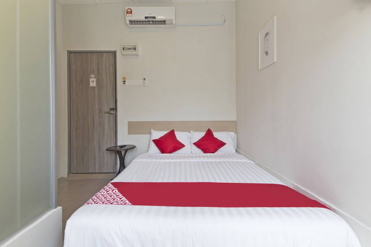 Una habitación de hotel con cama, una mesita de noche, una manta roja y un aire acondicionado instalado en la pared.