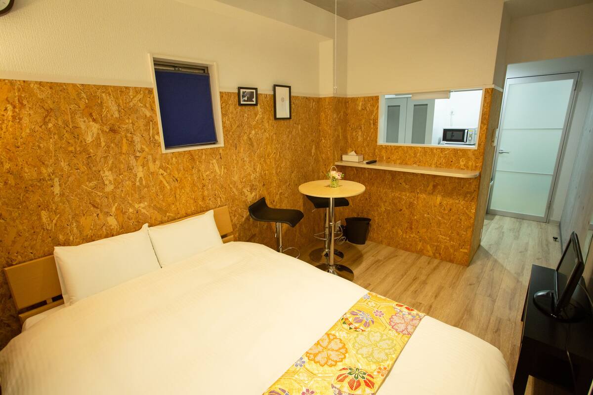 A hotel room with a bed, a small round table, two bar stools, a microwave, and a bathroom door.