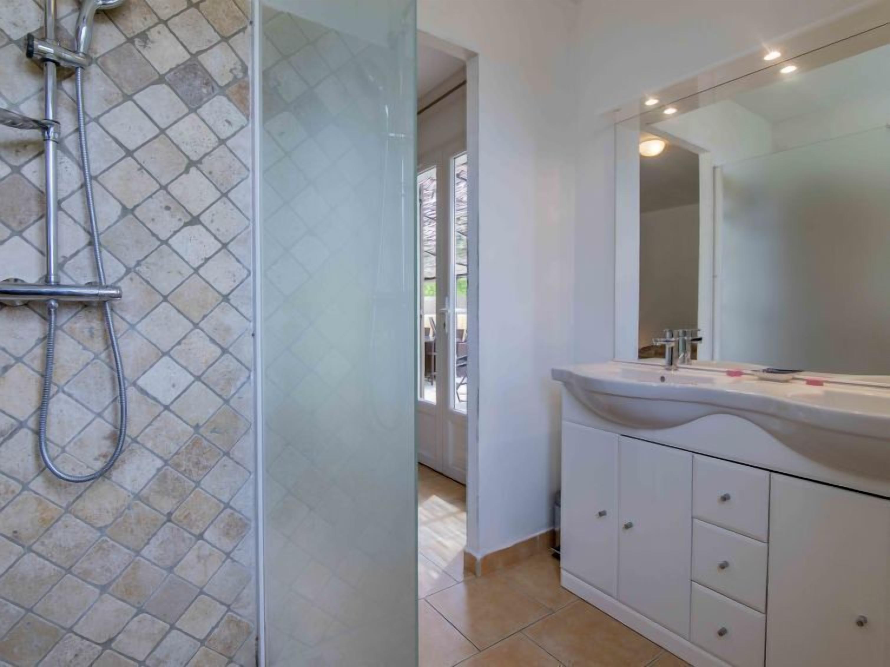 Une salle de bain avec une douche à parois de verre, un lavabo blanc avec un miroir et un sol carrelé.