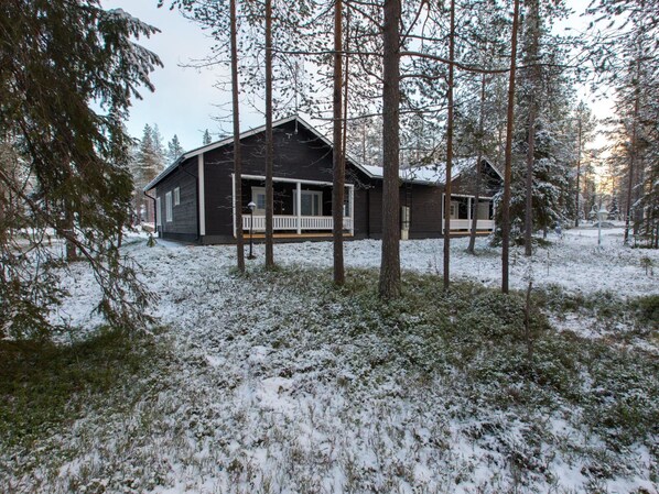 A two-story cabin with a porch, surrounded by tall trees and covered in snow.