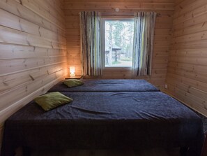 A room with wooden walls, two beds with dark bedding and green pillows, a bedside table with a lamp, and a window with striped curtains.