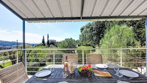 Área de refeições ao ar livre com uma mesa posta para quatro pessoas, uma garrafa de vinho e uma cesta de frutas.