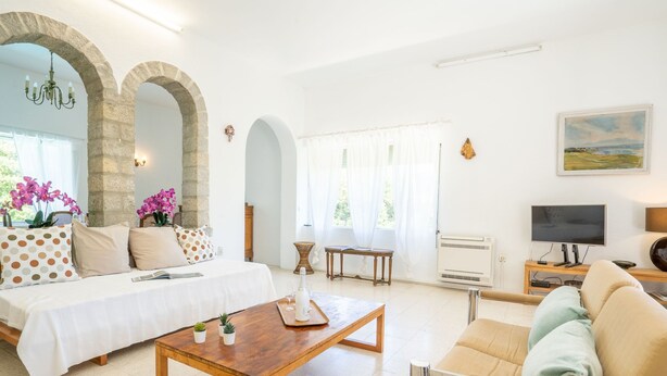 Uma sala de estar ampla com uma cama, um sofá, uma mesa de centro e uma TV. Apresenta arcos de pedra, um candelabro e um quadro na parede.
