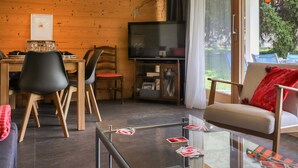 Un salón acogedor con paredes de madera, una zona de comedor con sillas, un televisor y una mesa de centro de vidrio con cartas.