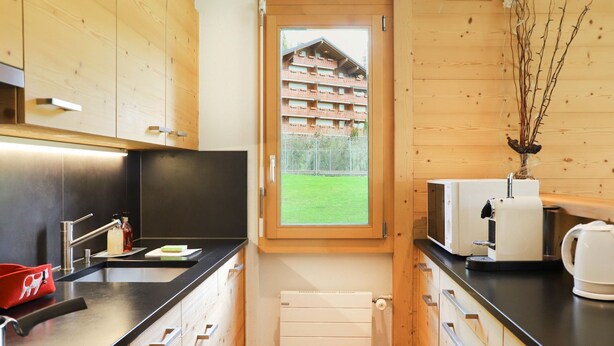 Cocina compacta con armarios de madera, horno integrado, cafetera y vista a un césped verde por una ventana.