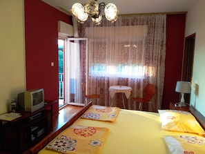 Ein Hotelzimmer mit Bett, Schreibtisch mit Fernseher, Sessel, kleinem Tisch und Fenster mit Vorhängen.