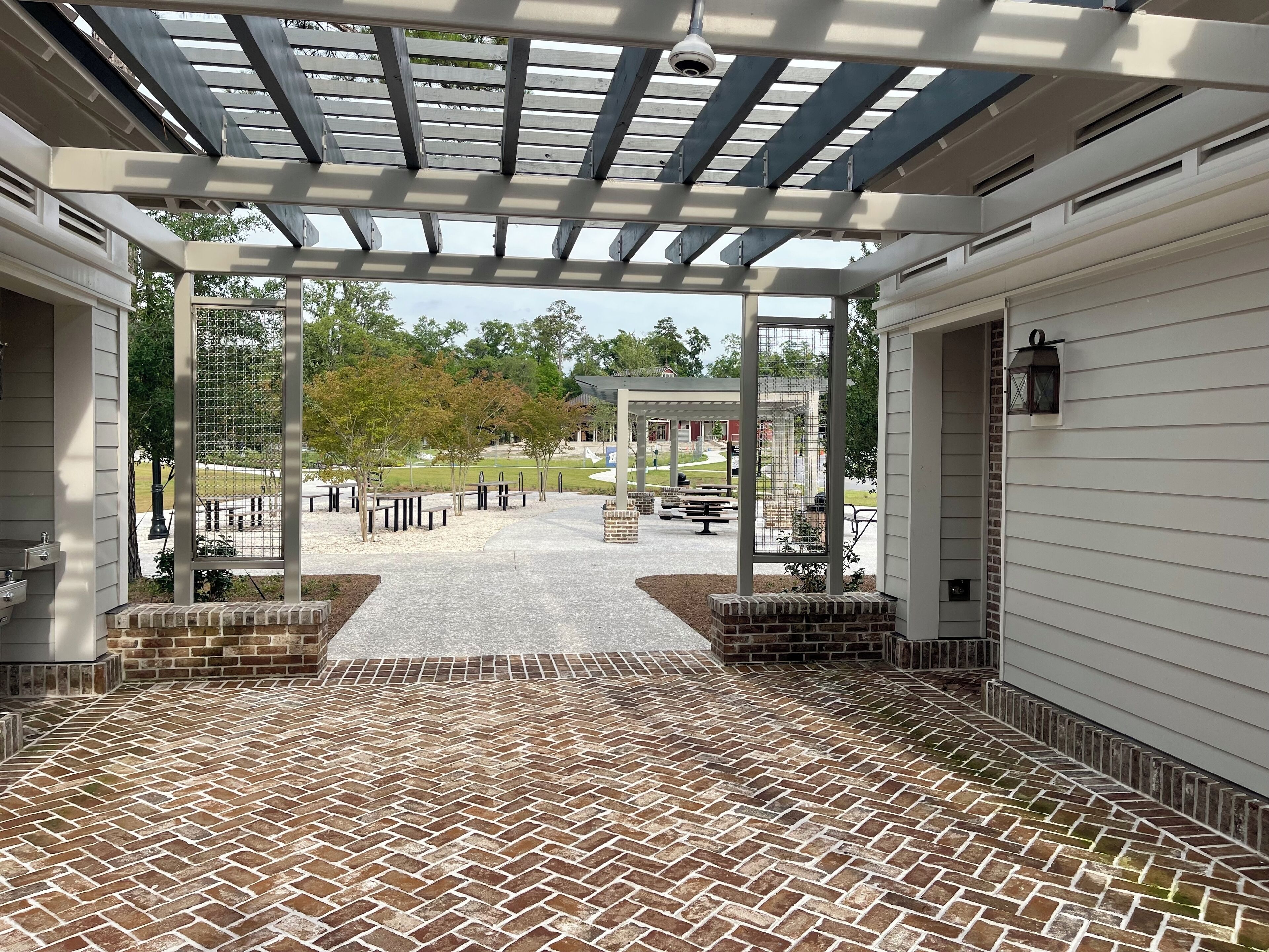 Un patio couvert avec un sol en briques et une vue sur un parc équipé de tables de pique-nique et d’un kiosque.