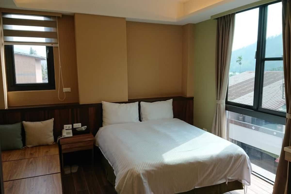 A hotel room with a large bed, a bedside table with a lamp, a window with curtains, and a view of the outside.
