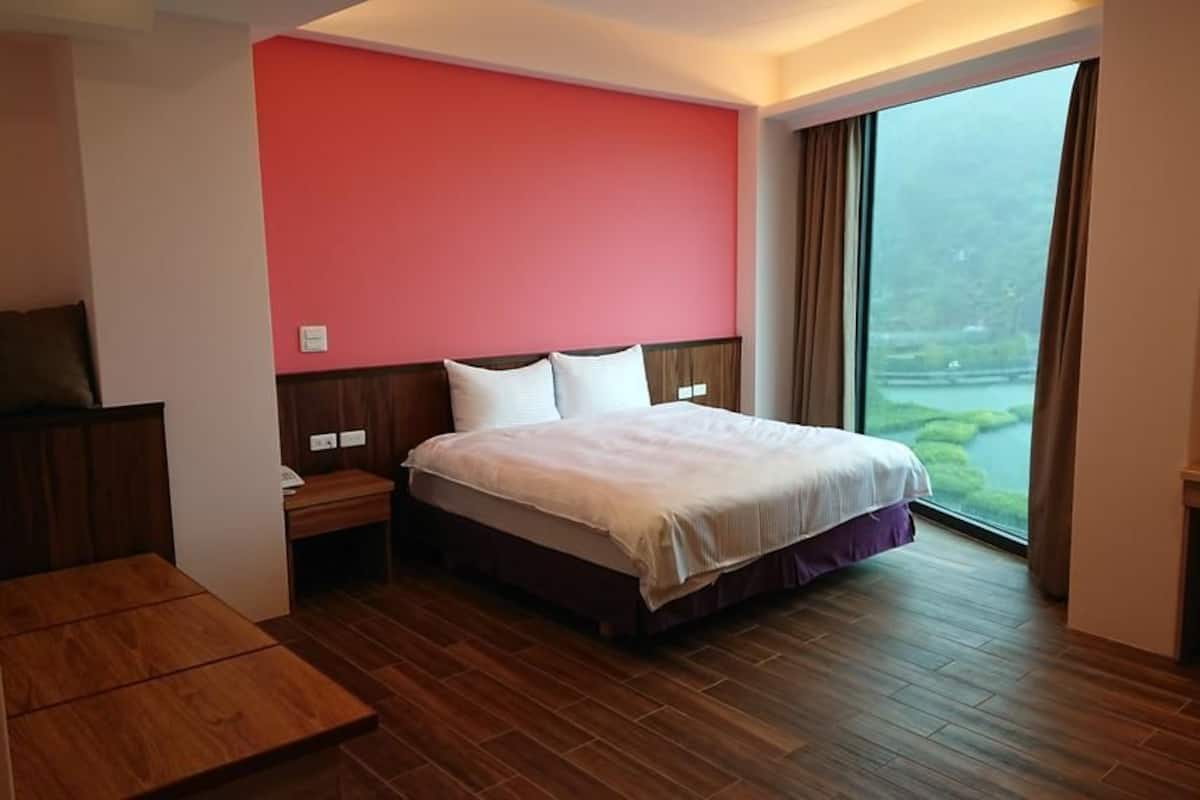 A hotel room with a large bed, a wooden desk, a TV, and a view of a river through a large window.