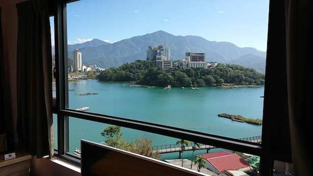 A room with a large window offering a view of a lake, buildings, and mountains.