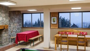 A cozy room with a pool table, a wooden bench, and a table with a red tablecloth. There are large windows offering a view of trees and a cabin.