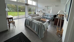 Amplia sala de estar con un sofá grande, una mesa de centro y un comedor con mesa y sillas. Amplias ventanas ofrecen luz natural y vistas al exterior.