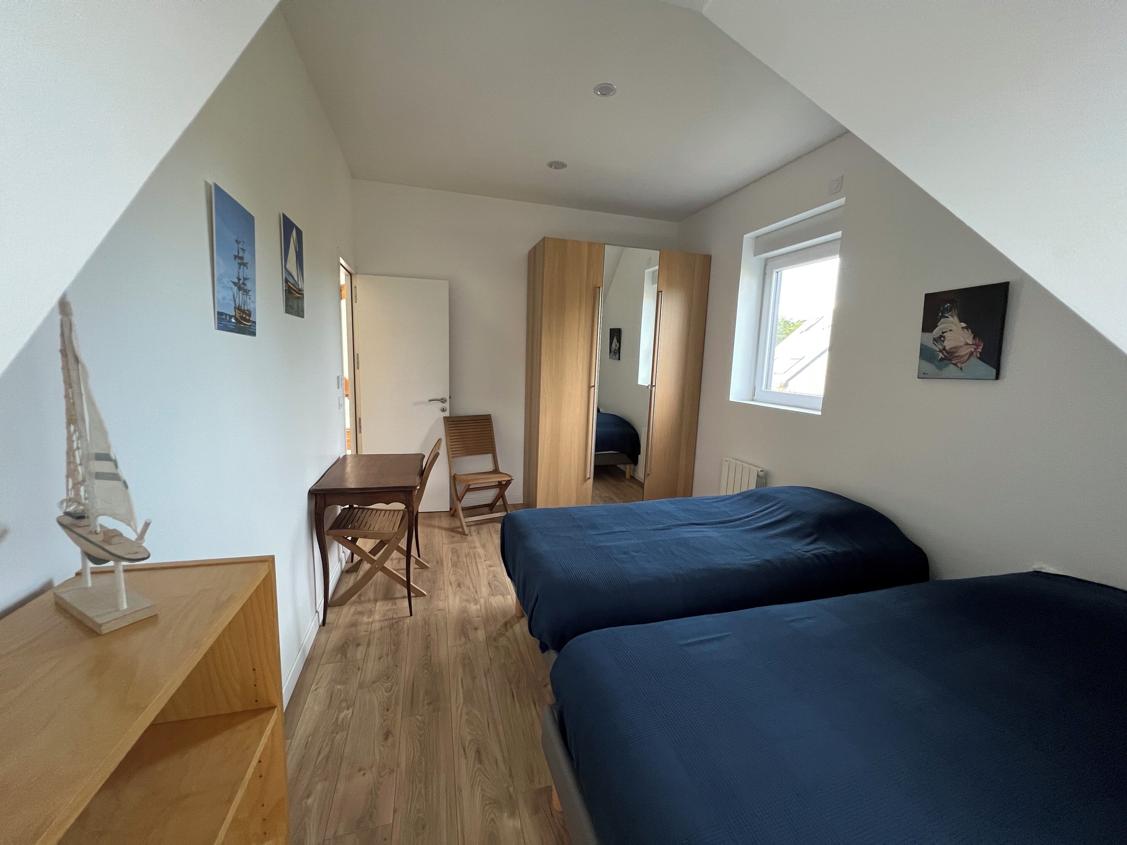 A small, triangular room with two beds, a wooden desk, a chair, and a small table with a model ship.