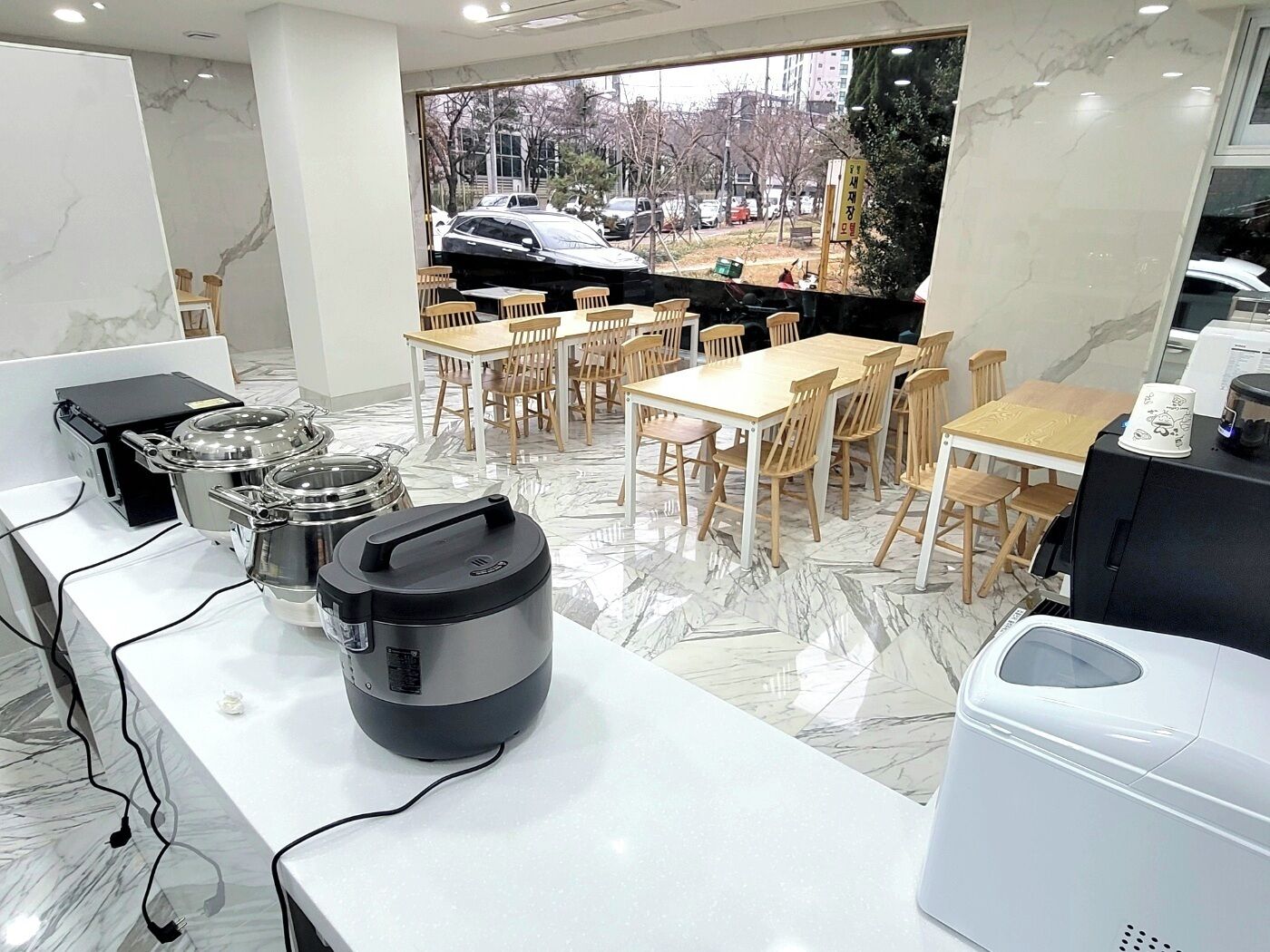 Un espace repas moderne avec un sol en marbre, des tables et des chaises en bois, ainsi que des appareils de cuisine comme une machine à café et un cuiseur à riz.
