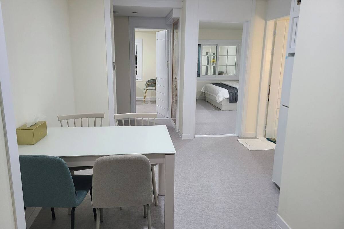 A dining area with a white table and chairs, a yellow tissue box, and a view into a bedroom with a bed and a window.