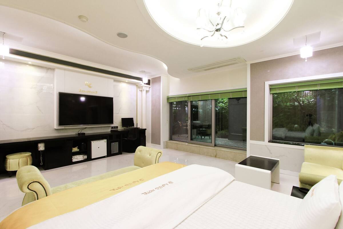 A modern hotel room with a large bed, a flat-screen TV, a desk with a computer, and a view of greenery through the windows.