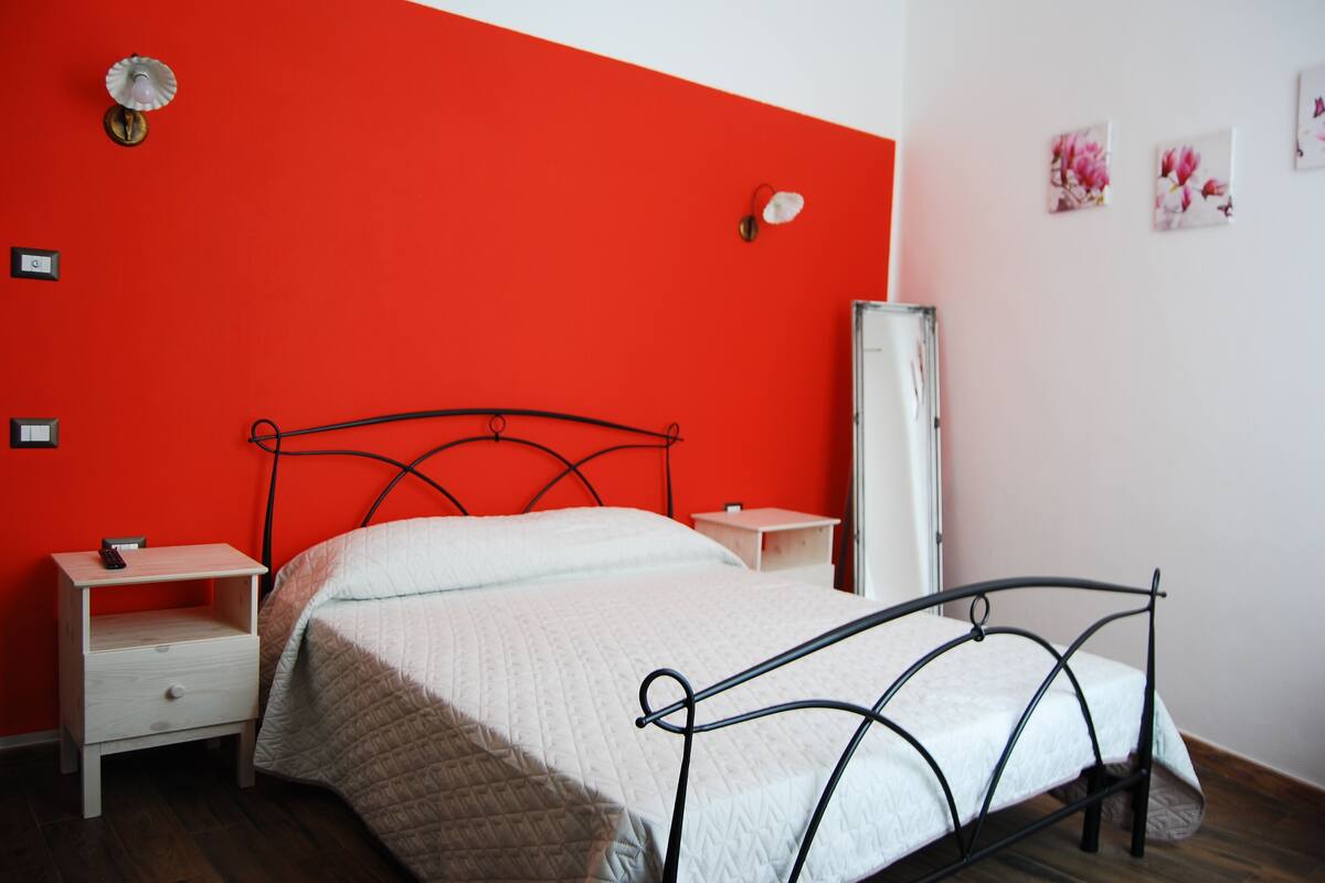 A bedroom with a bed, nightstands, a mirror, and a red accent wall.