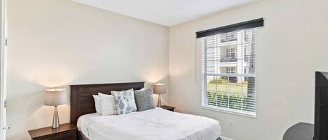 Un dormitorio con cama, mesitas de noche, lámpara, ventana con persianas y televisor.