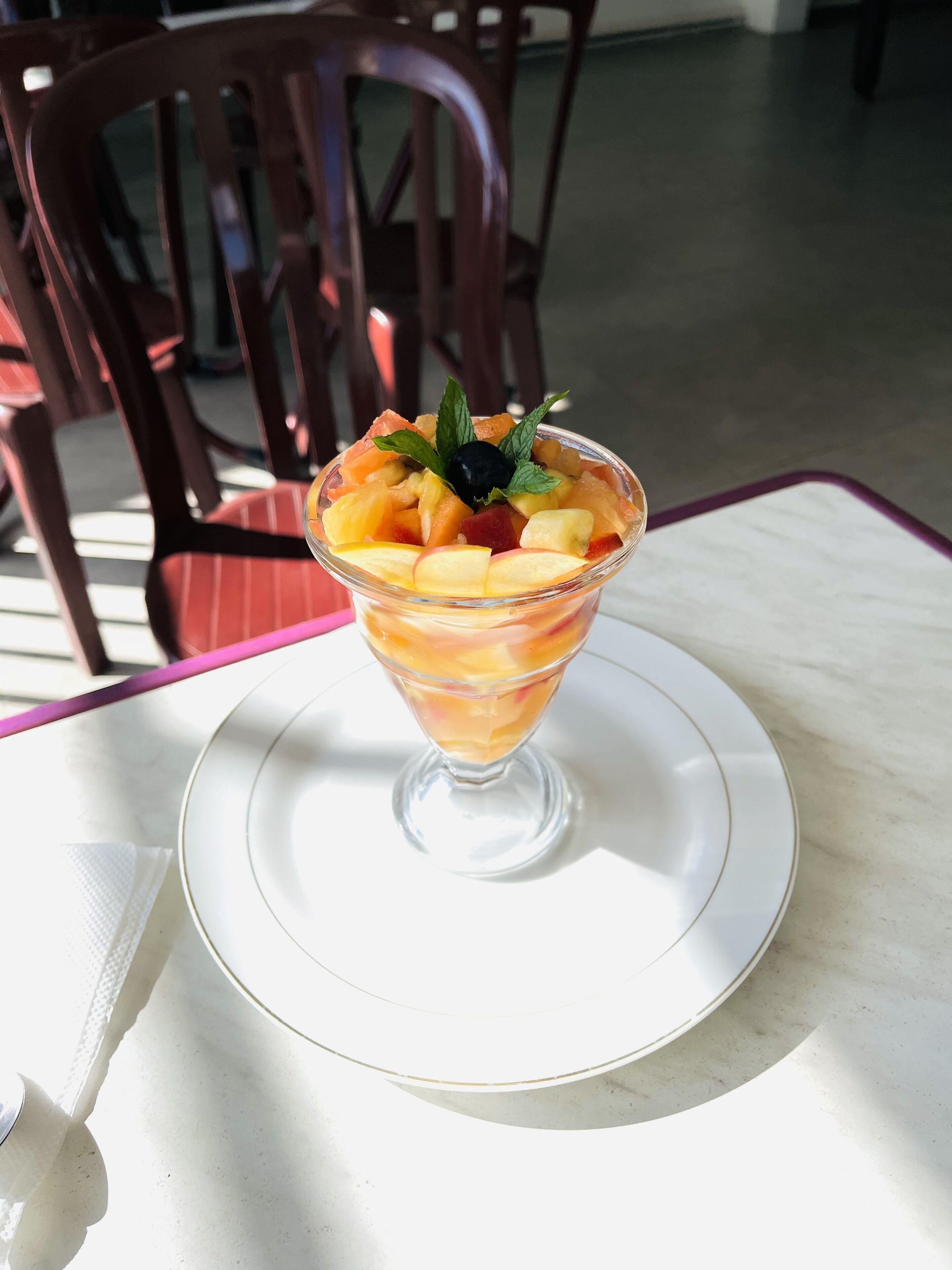 Een fruitsalade in een glazen schaal op een wit bord, met een servet en een lepel ernaast.