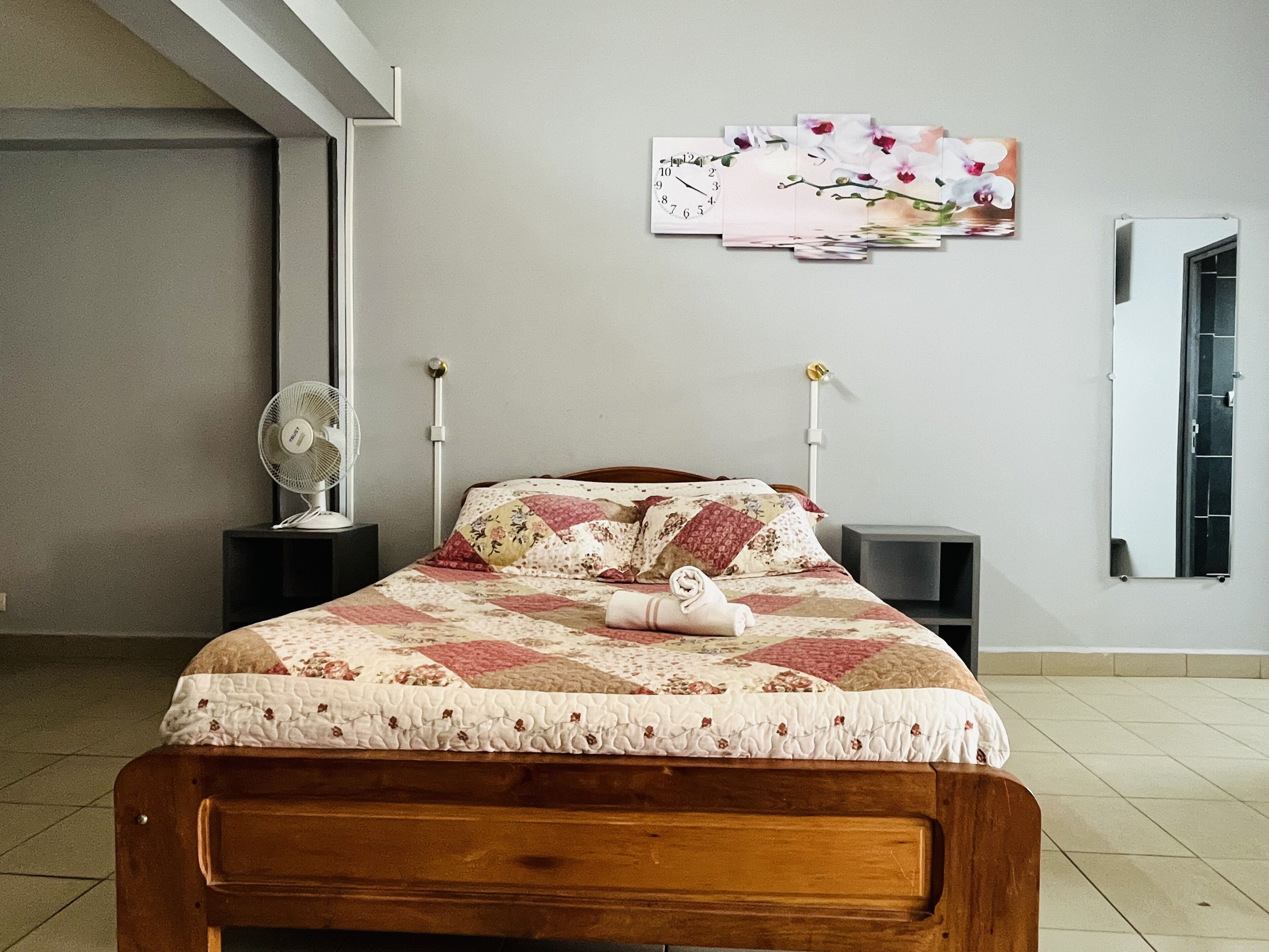 Un dormitorio con cama de madera, colcha, reloj de pared, ventilador y espejo.
