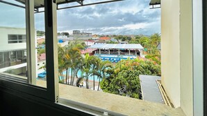 A view from a window overlooking a swimming pool area with a covered seating area, surrounded by greenery and buildings. - Hotel Appartement meublé Les Flots Bleu (Antananarivo)
