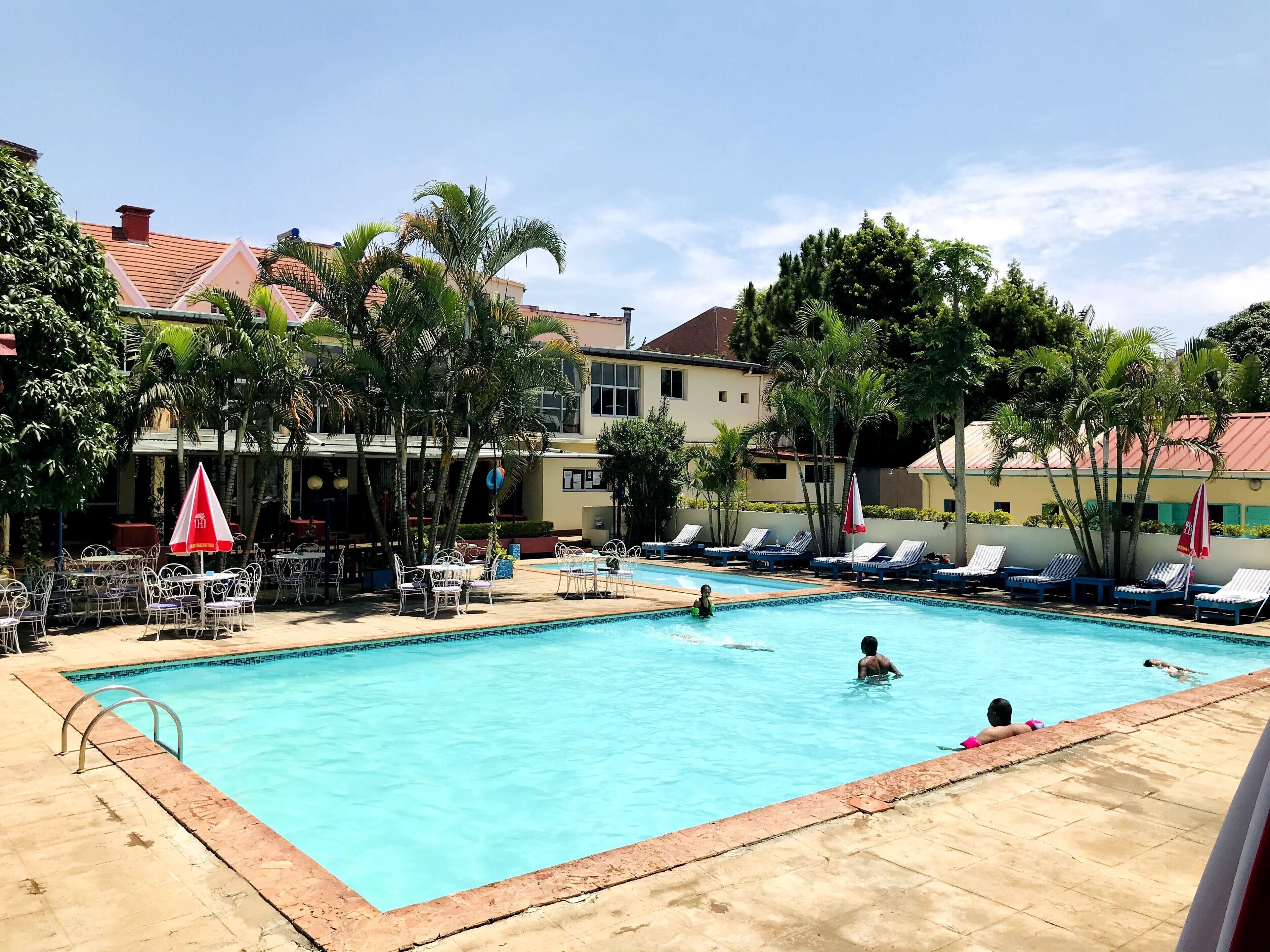 A swimming pool with lounge chairs and umbrellas, surrounded by palm trees and buildings.