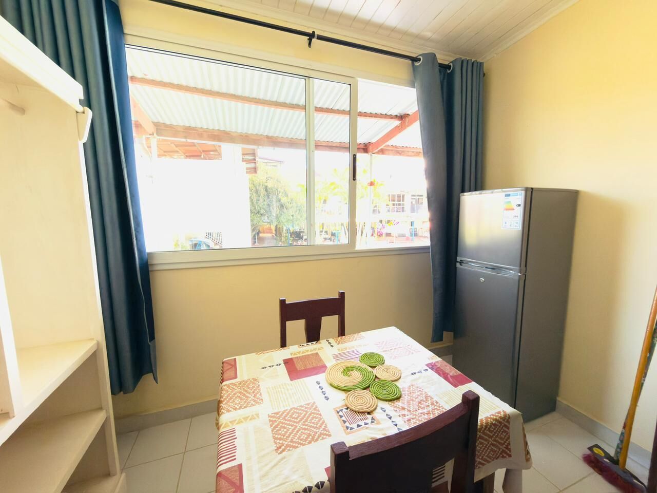 Una habitación pequeña con una mesa dispuesta para dos personas, un refrigerador y una ventana con cortinas.