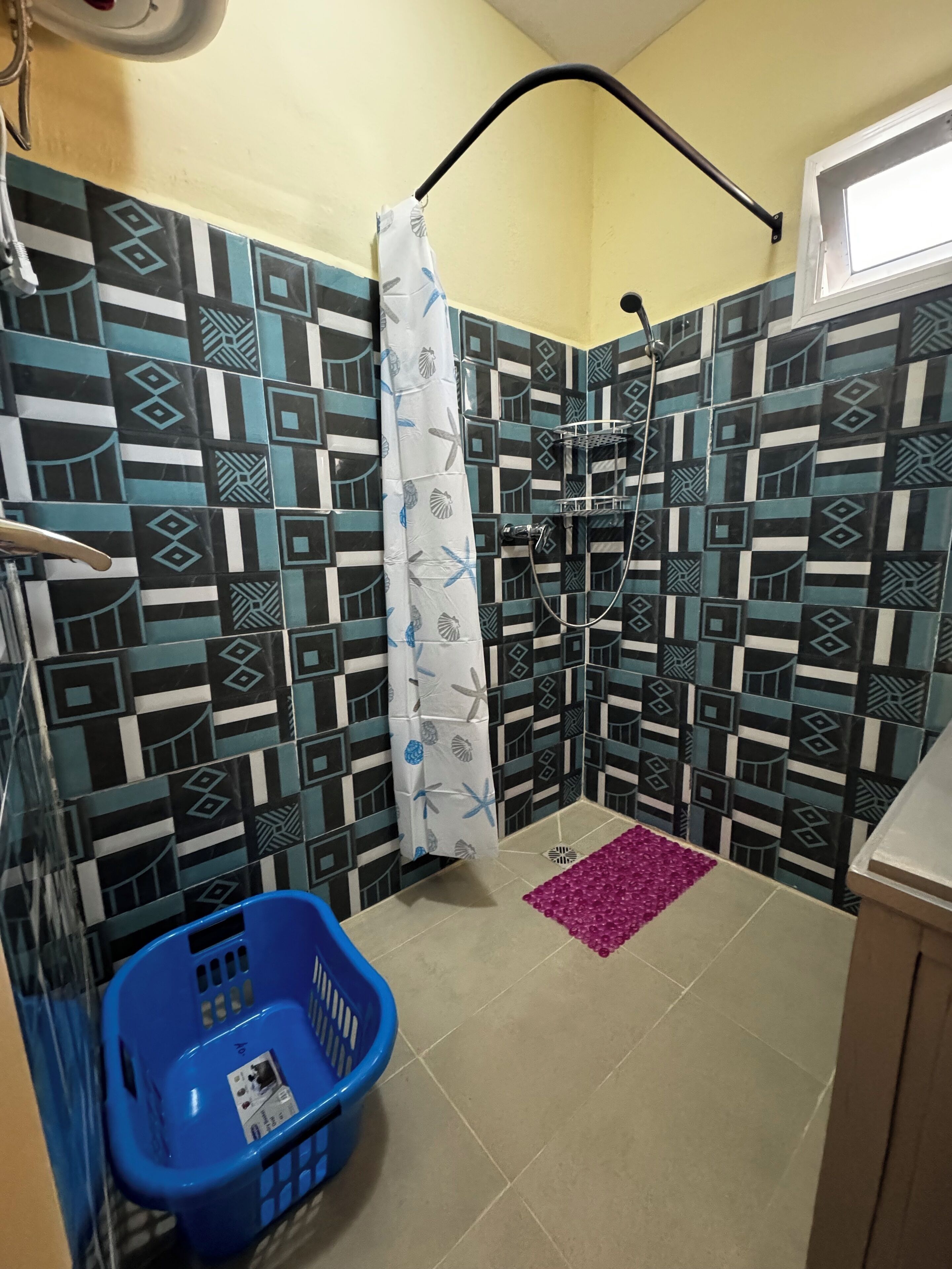 Un baño con baldosas estampadas, una ducha, una cesta de plástico azul y un tapete rosa.