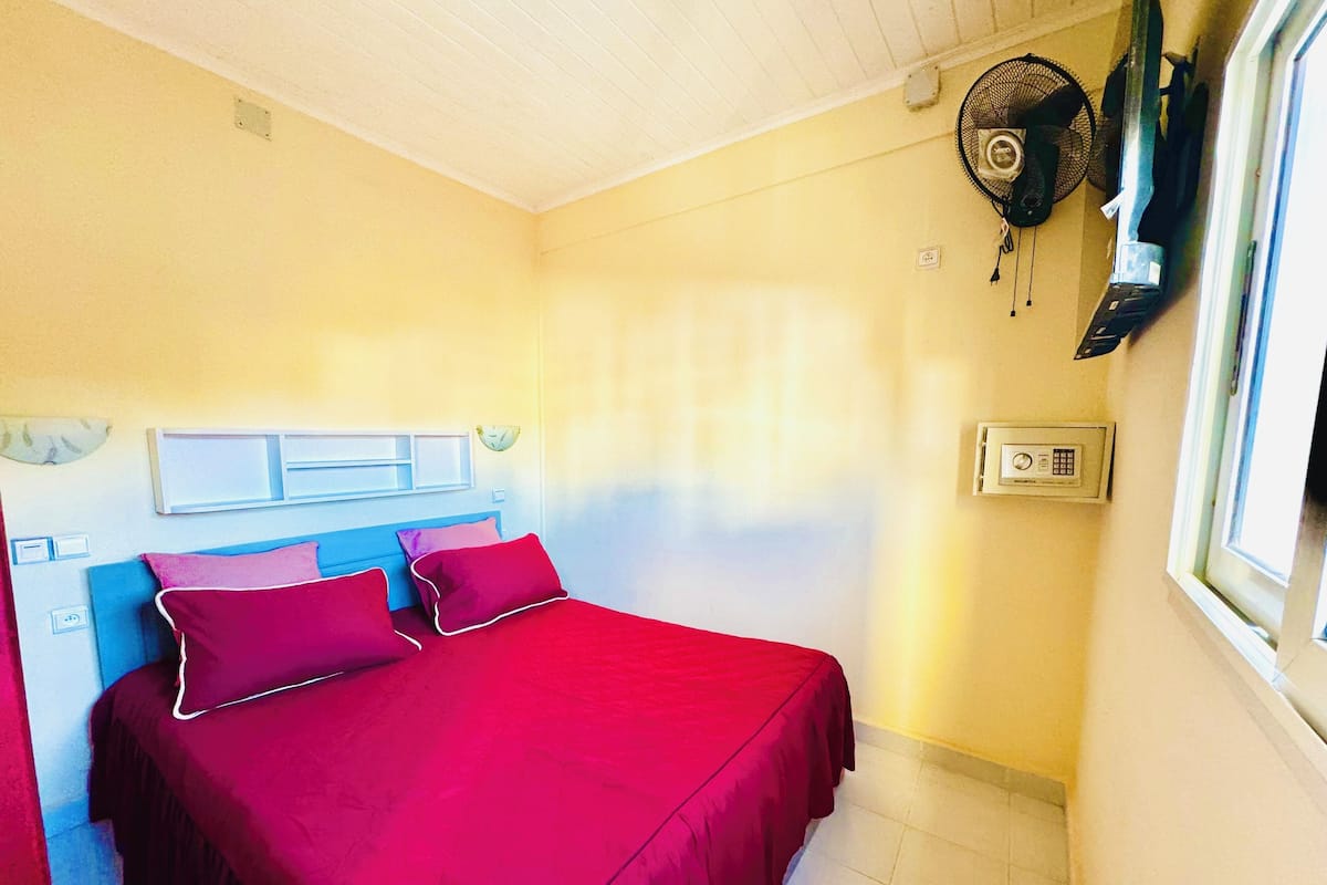 Una habitación con una cama, un ventilador de pared y una ventana.