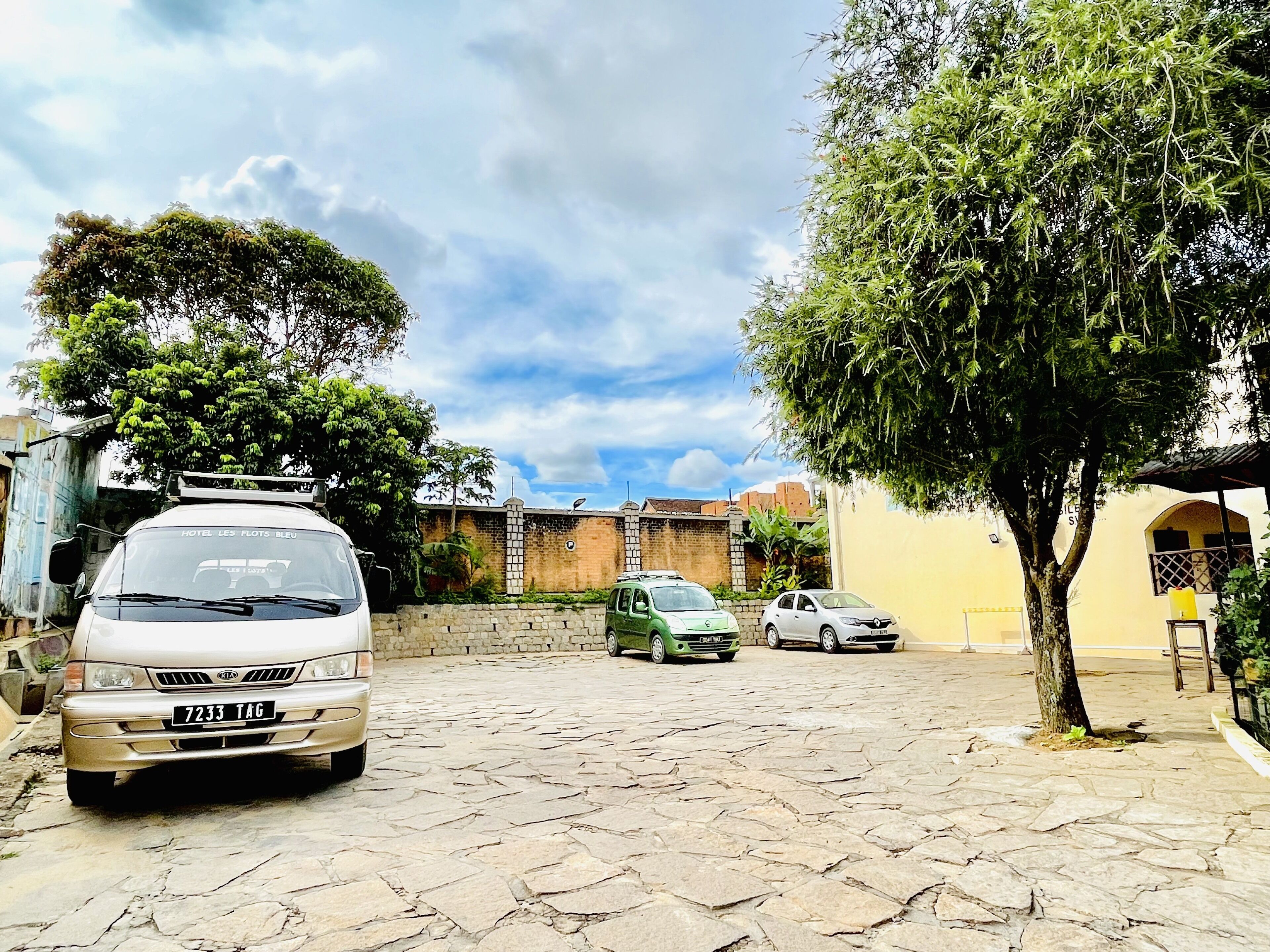 Un patio empedrado con una camioneta blanca, una camioneta verde y un coche plateado estacionados. Se ven árboles y un muro de ladrillo al fondo.