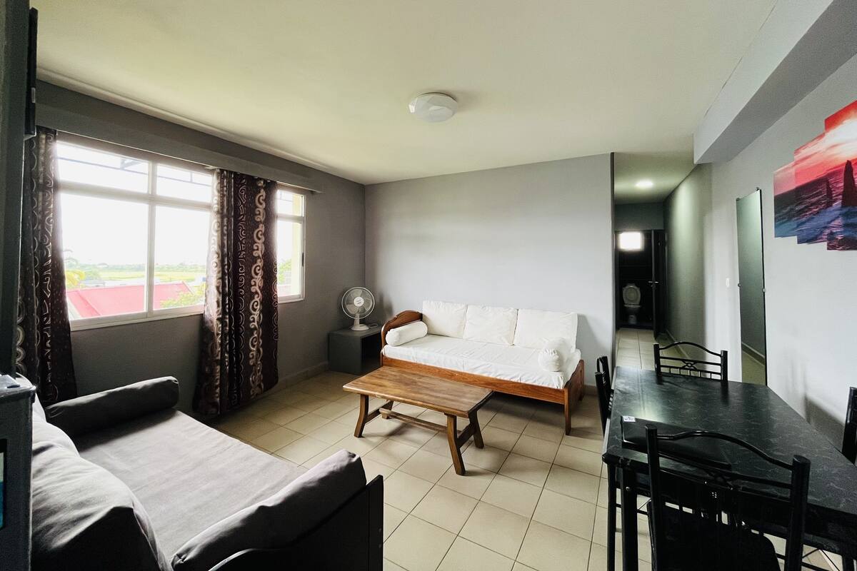 Una habitación con cama, sofá, mesa de comedor con sillas, ventilador y ventana con cortinas.