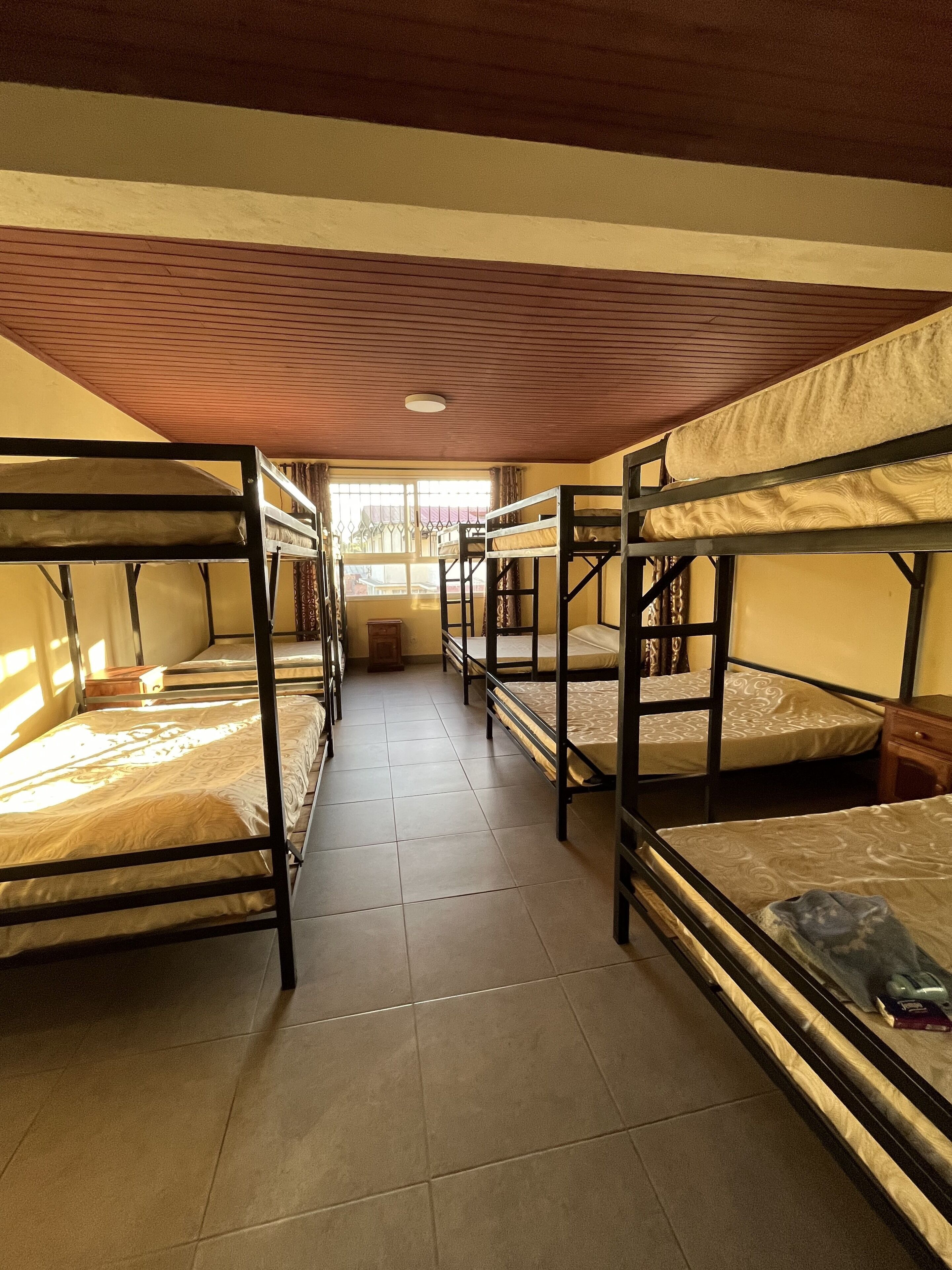 A dormitory room with bunk beds arranged in two rows, a tiled floor, and a window with curtains.