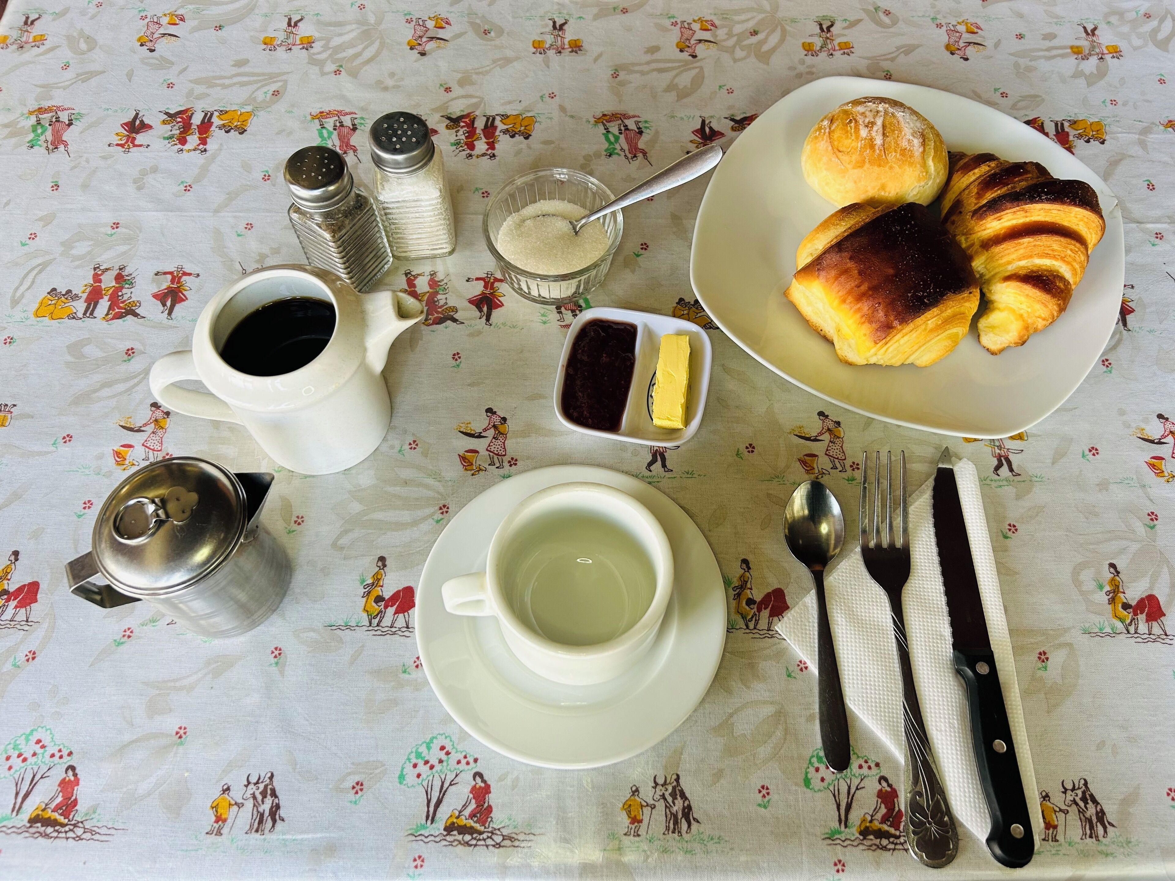A table with a floral tablecloth, a plate of croissants, a cup of coffee, a teapot, salt and pepper shakers, a butter dish, and cutlery.