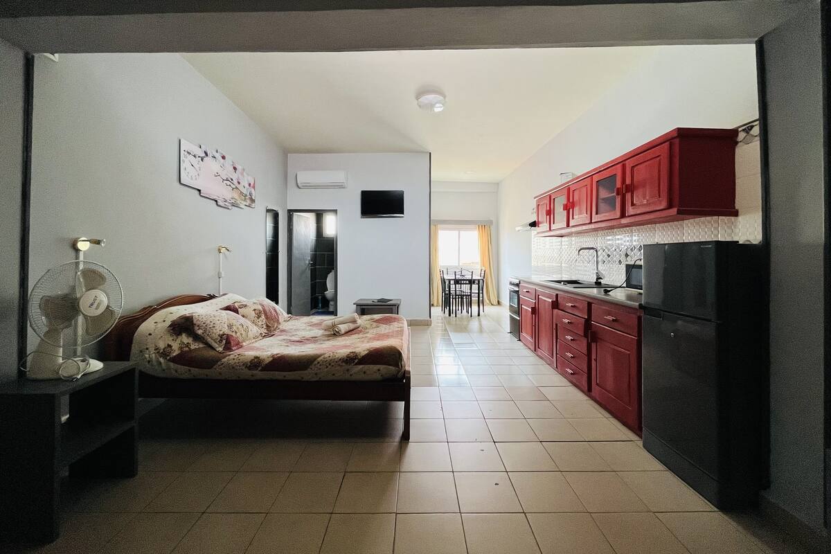 Habitación de hotel compacta con cama, mesita de noche y un pequeño escritorio. Cuenta con una kitchenette con armarios rojos y un refrigerador negro. Un televisor de pantalla plana está montado en la pared.