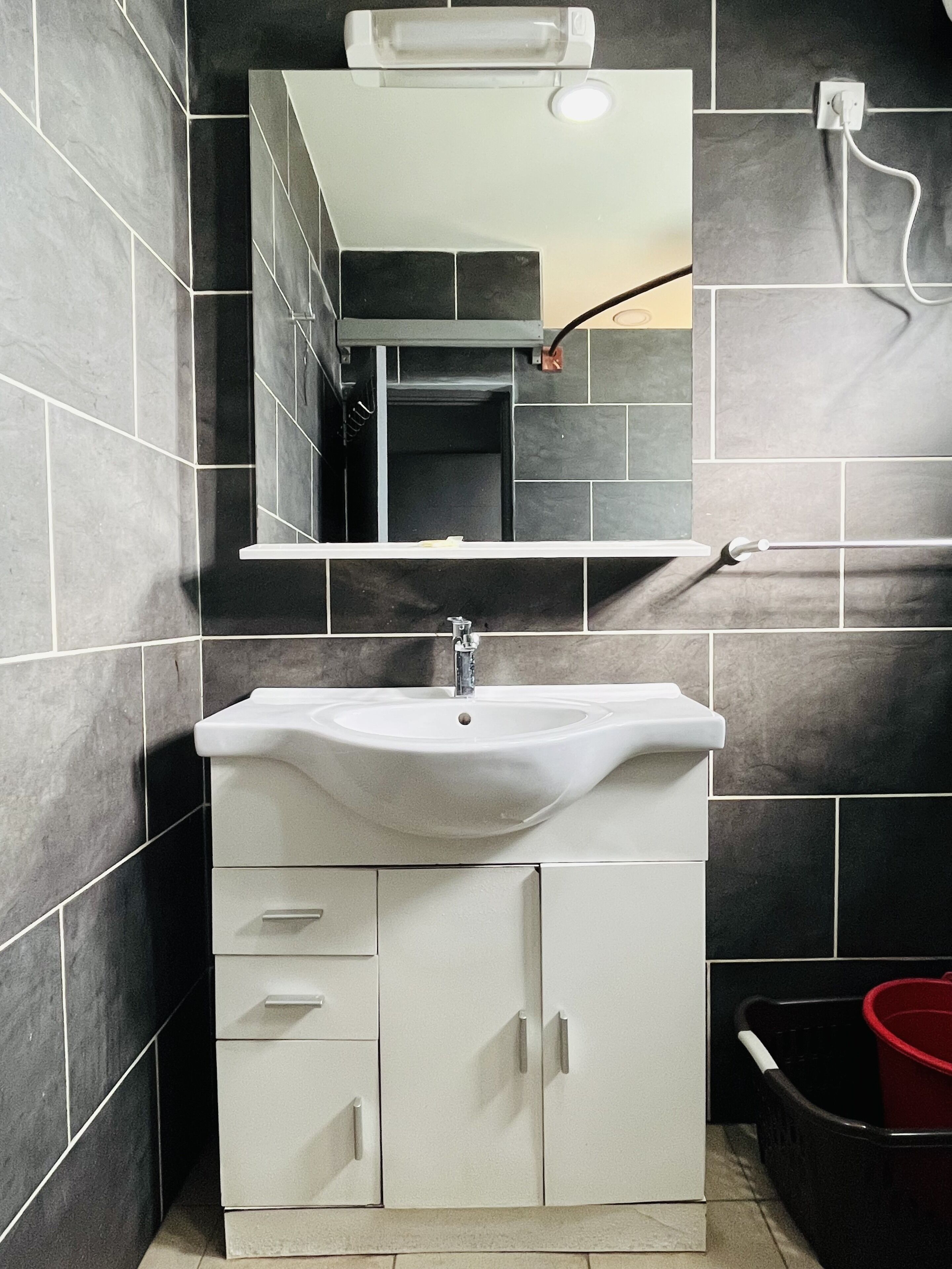 Un baño con un lavabo blanco, un espejo encima y paredes cubiertas de baldosas oscuras.
