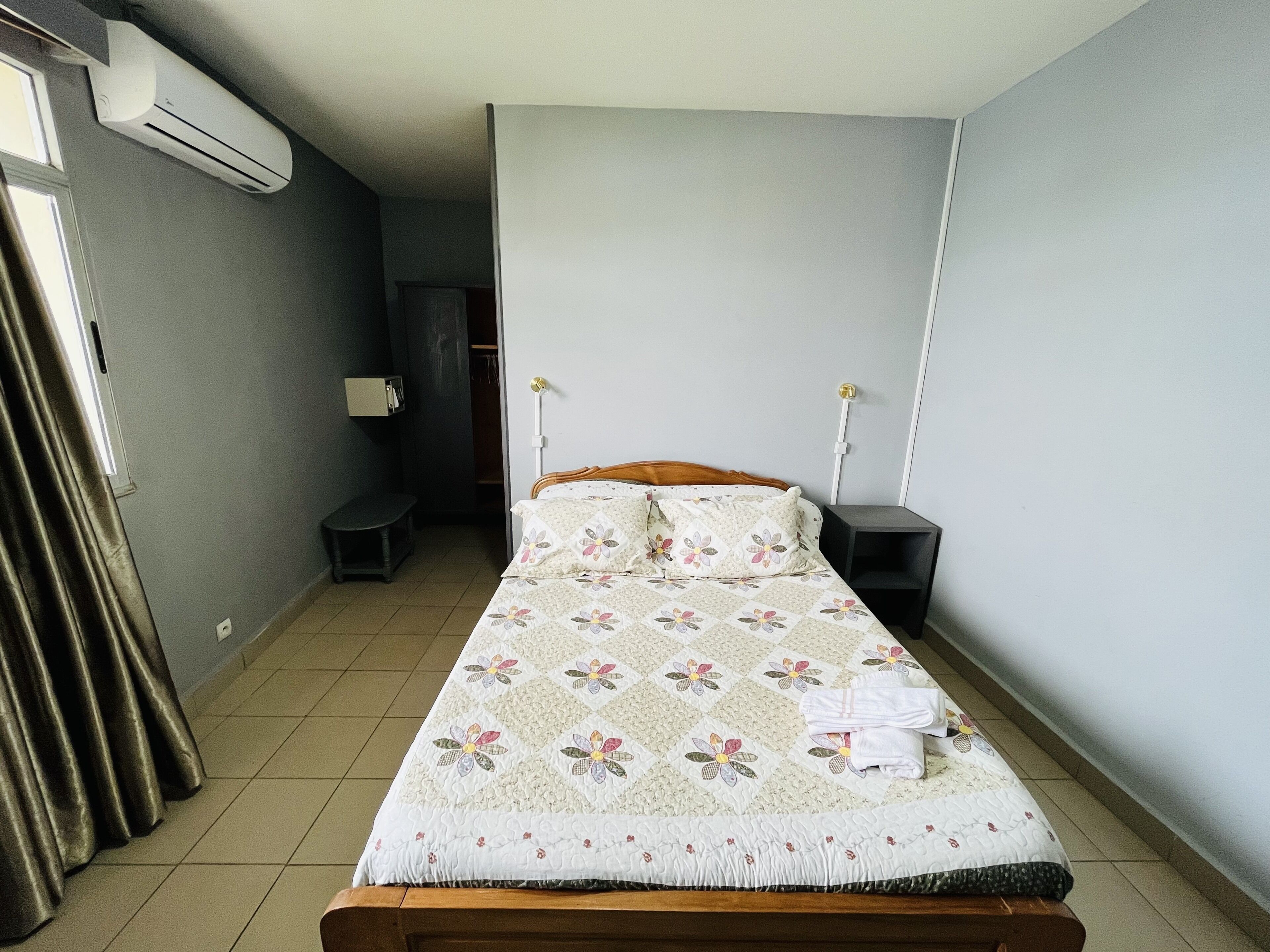 Ein Einzelbett mit floralem Überwurf, ein Nachttisch, ein Fenster mit Vorhängen und eine Klimaanlage an der Wand.