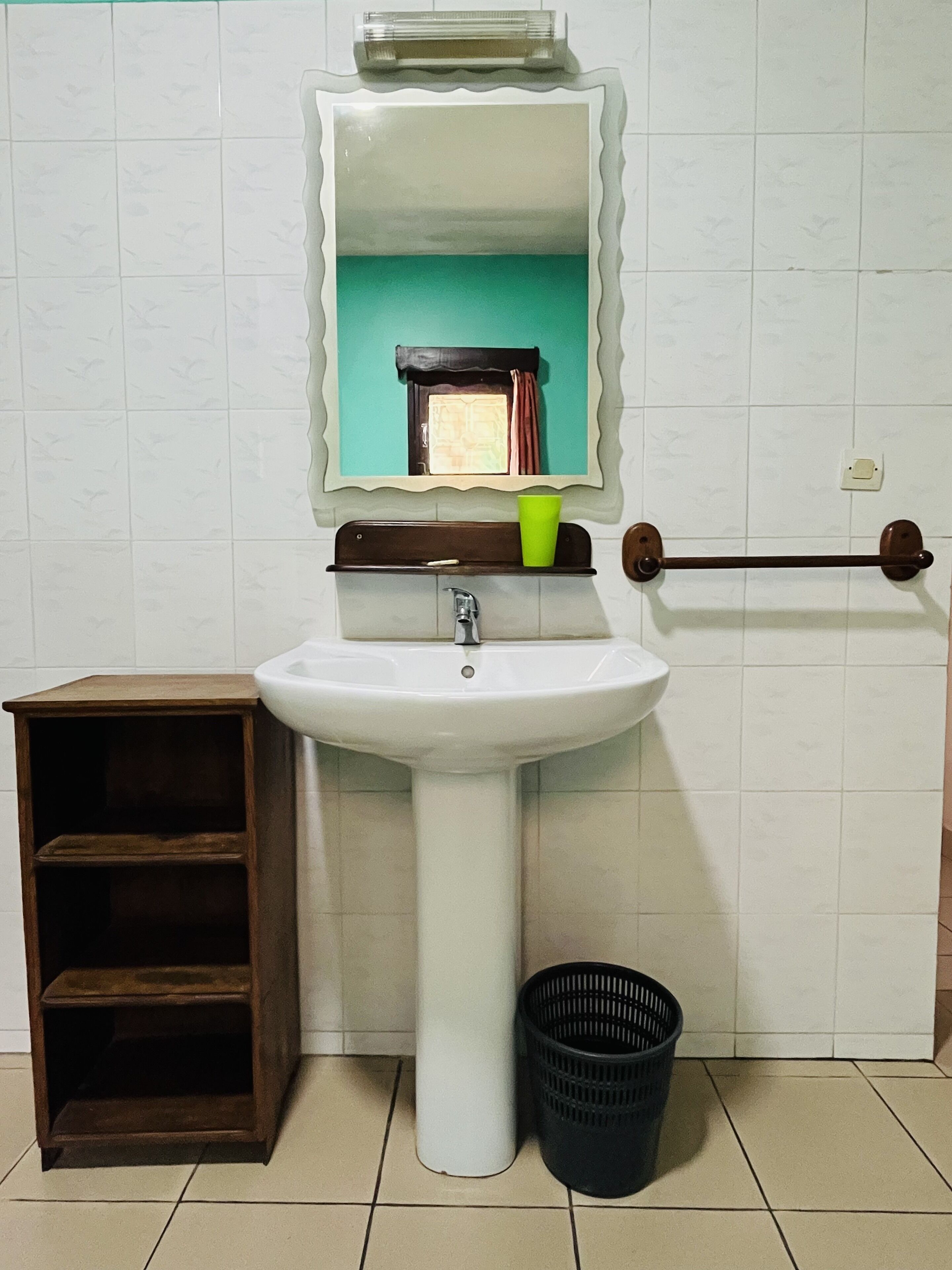 A bathroom with a white sink, a mirror, a shelf with a green cup, a wooden shelf unit, and a black trash bin.